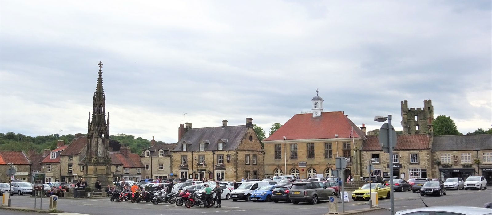 A wide view of Helmsley Market Place: the Feversham Monument, the limestone town hall and shops, and the ruins of Helmsley Castle on the right